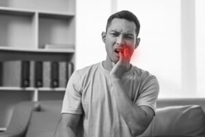 A man sits on a sofa, grimacing in pain and holding his jaw, with a red highlight indicating wisdom tooth or molar pain, emphasizing dental discomfort.