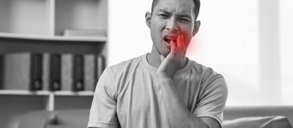 A man sits on a sofa, grimacing in pain and holding his jaw, with a red highlight indicating wisdom tooth or molar pain, emphasizing dental discomfort.